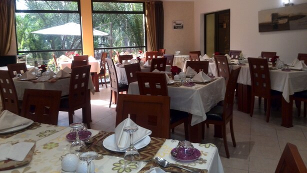 Un espace repas avec des tables disposées pour accueillir plusieurs convives, de grandes fenêtres avec des rideaux et une vue sur la verdure environnante.