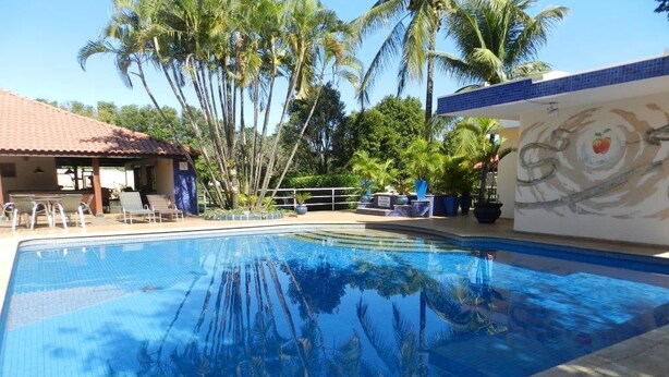 A clear swimming pool with lounge chairs and a covered seating area, surrounded by palm trees and a building with a tiled roof. - Paradise Inn Hotel e Lazer (Catanduva)