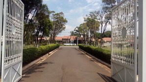 A gated entrance to a property with a paved driveway, surrounded by trees and a building with a red roof in the background. - Paradise Inn Hotel e Lazer (Catanduva)