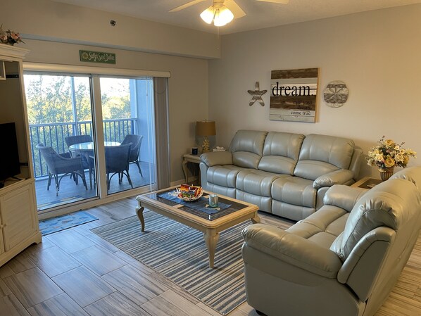 A living room with a grey leather sofa, a coffee table, and a TV. There is a sliding glass door leading to an outdoor area with a patio table and chairs.