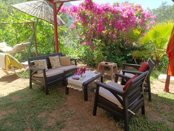 Un espace extérieur aménagé avec un canapé, des fauteuils et une table, le tout entouré d’une végétation luxuriante et de fleurs aux couleurs vives.