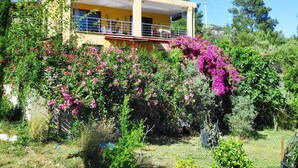 Un edificio de dos pisos con balcón, rodeado de abundante vegetación y plantas en flor.