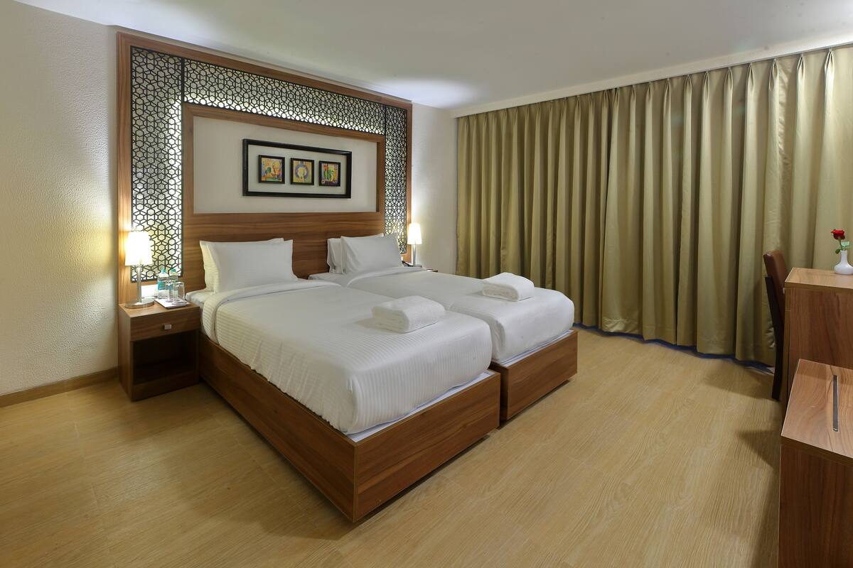 A hotel room with a double bed, wooden headboard, a desk with a chair, a TV, and a large window with curtains.