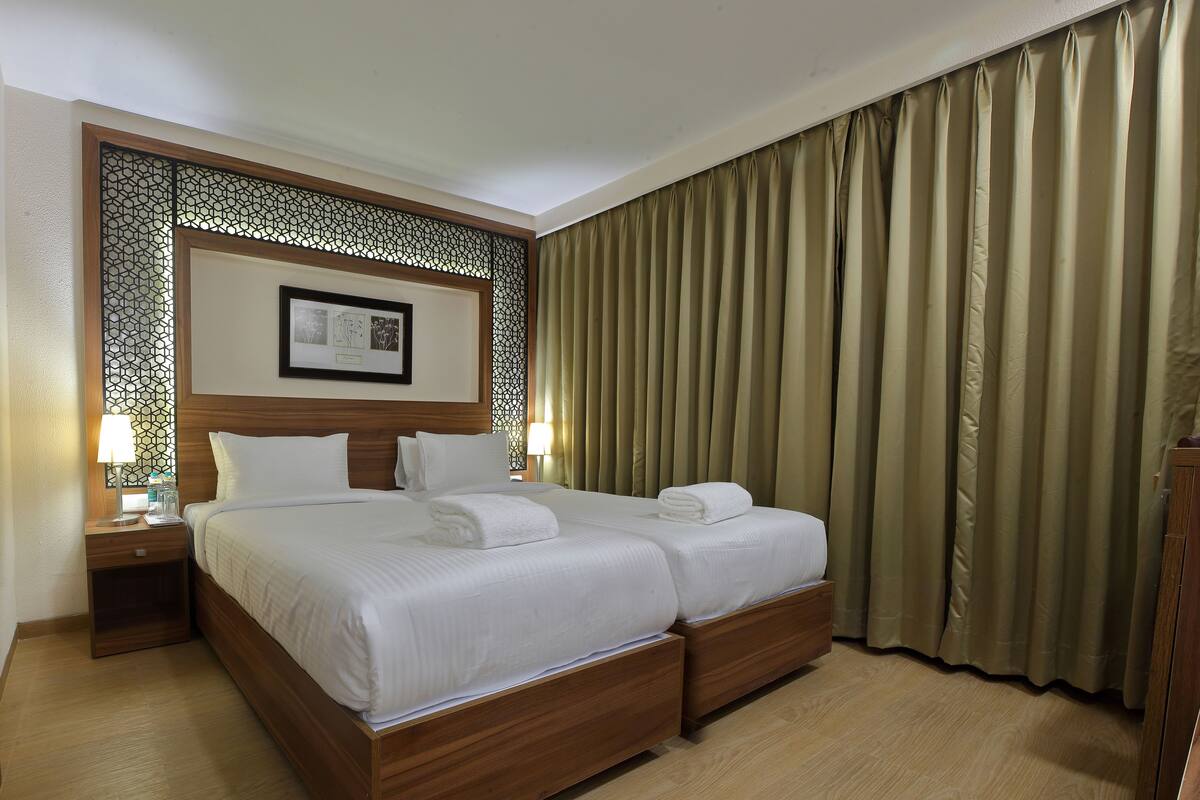 A hotel room with a double bed, two bedside tables with lamps, a decorative headboard, and a large window with curtains.