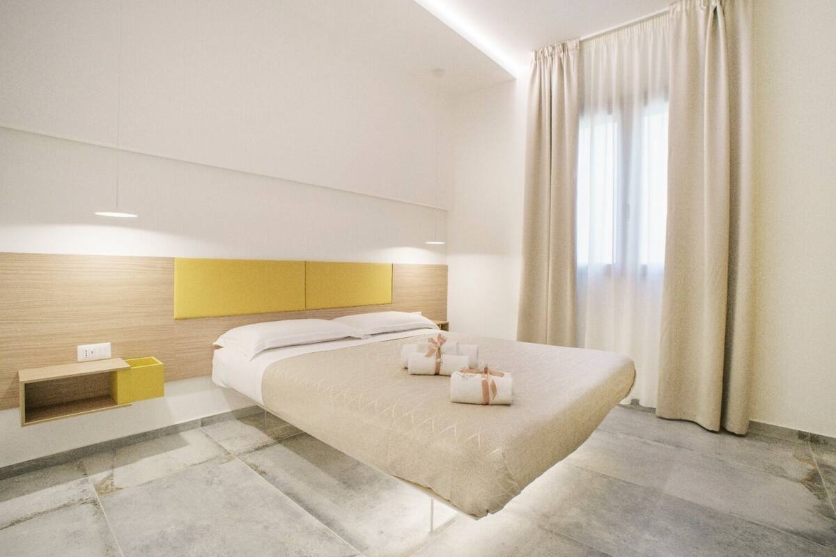 A modern hotel room with a large bed, a wooden headboard, a bedside table with a yellow box, and a large window with curtains.