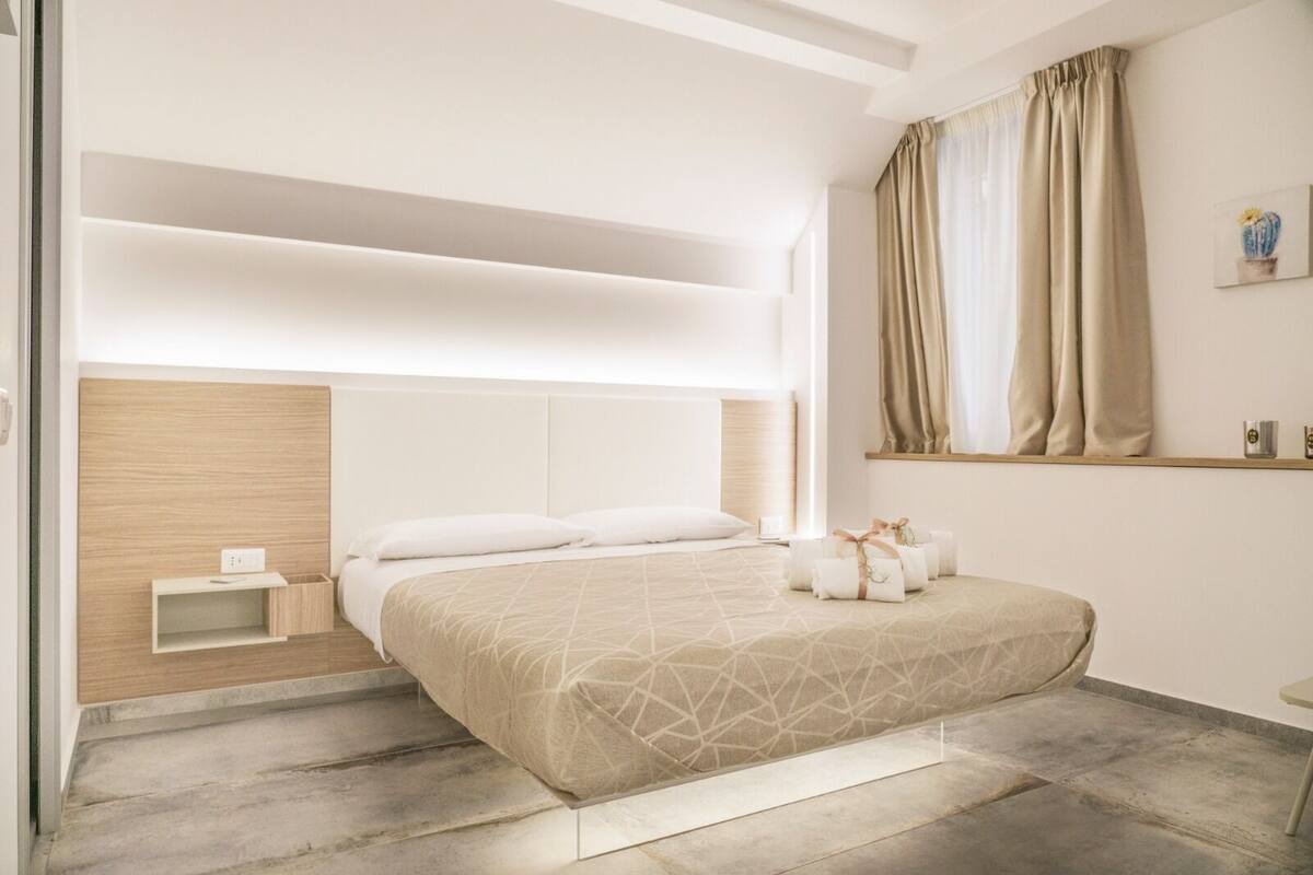 A modern bedroom with a large bed, a bedside table, a built-in shelf, and a window with curtains.