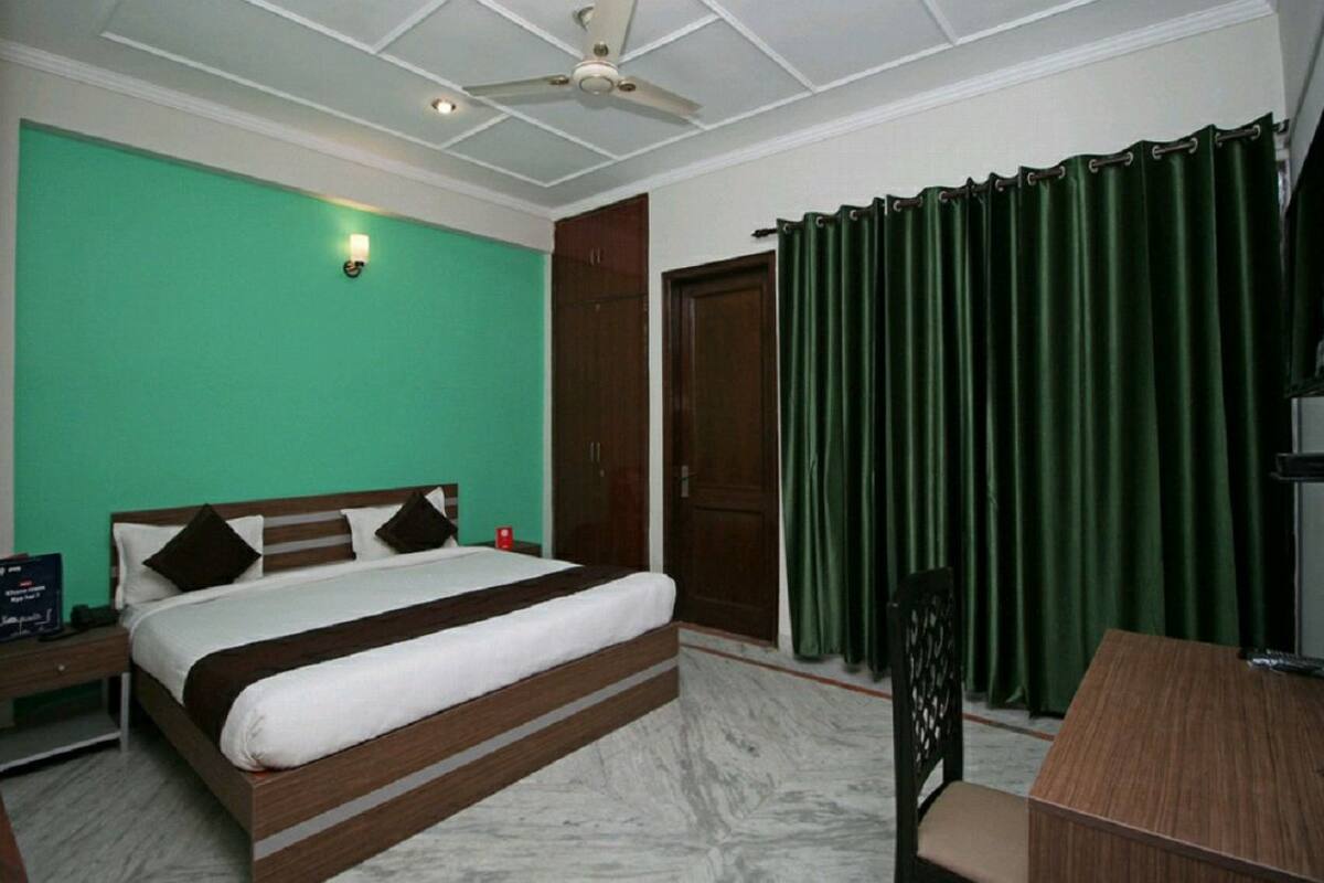 A hotel room with a bed, a desk, a chair, a ceiling fan, and a green curtain.