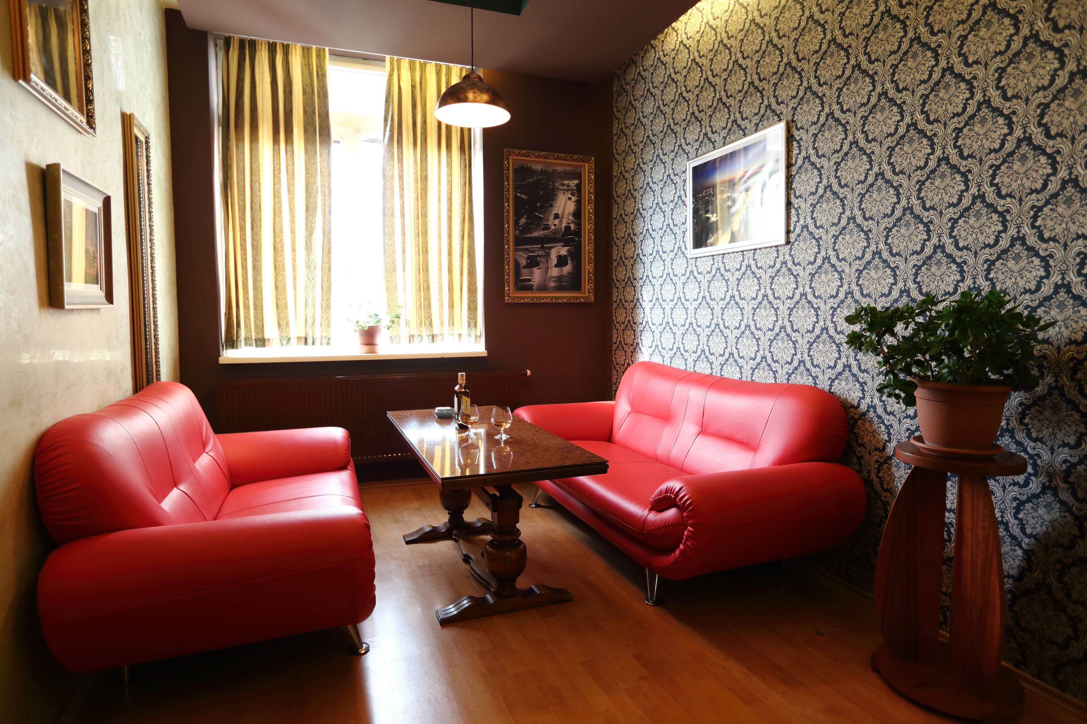 Ein Zimmer mit zwei roten Ledersofas, einem Holztisch mit Flasche und Gläsern, einer Topfpflanze und Wandpapier mit Muster.
