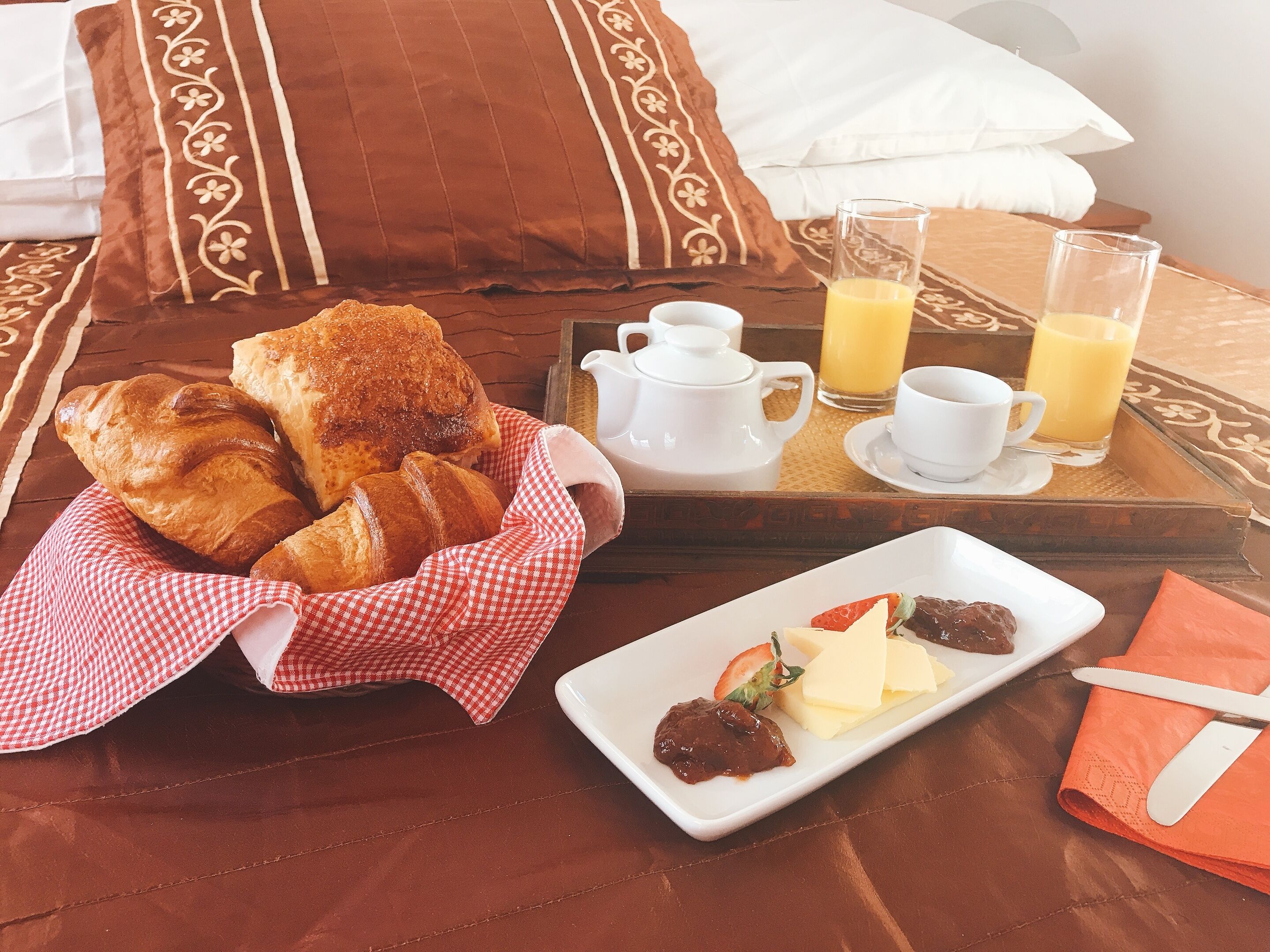 Ein Frühstücksbrett mit Croissants, einem Teller mit Erdbeere und Käse, einer Tasse Kaffee und einem Glas Orangensaft auf einem Bett.
