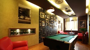 Une table de billard dans une pièce où se trouvent des chaises rouges, un porte-billes et un mur décoré de tableaux encadrés et d’un tableau noir.