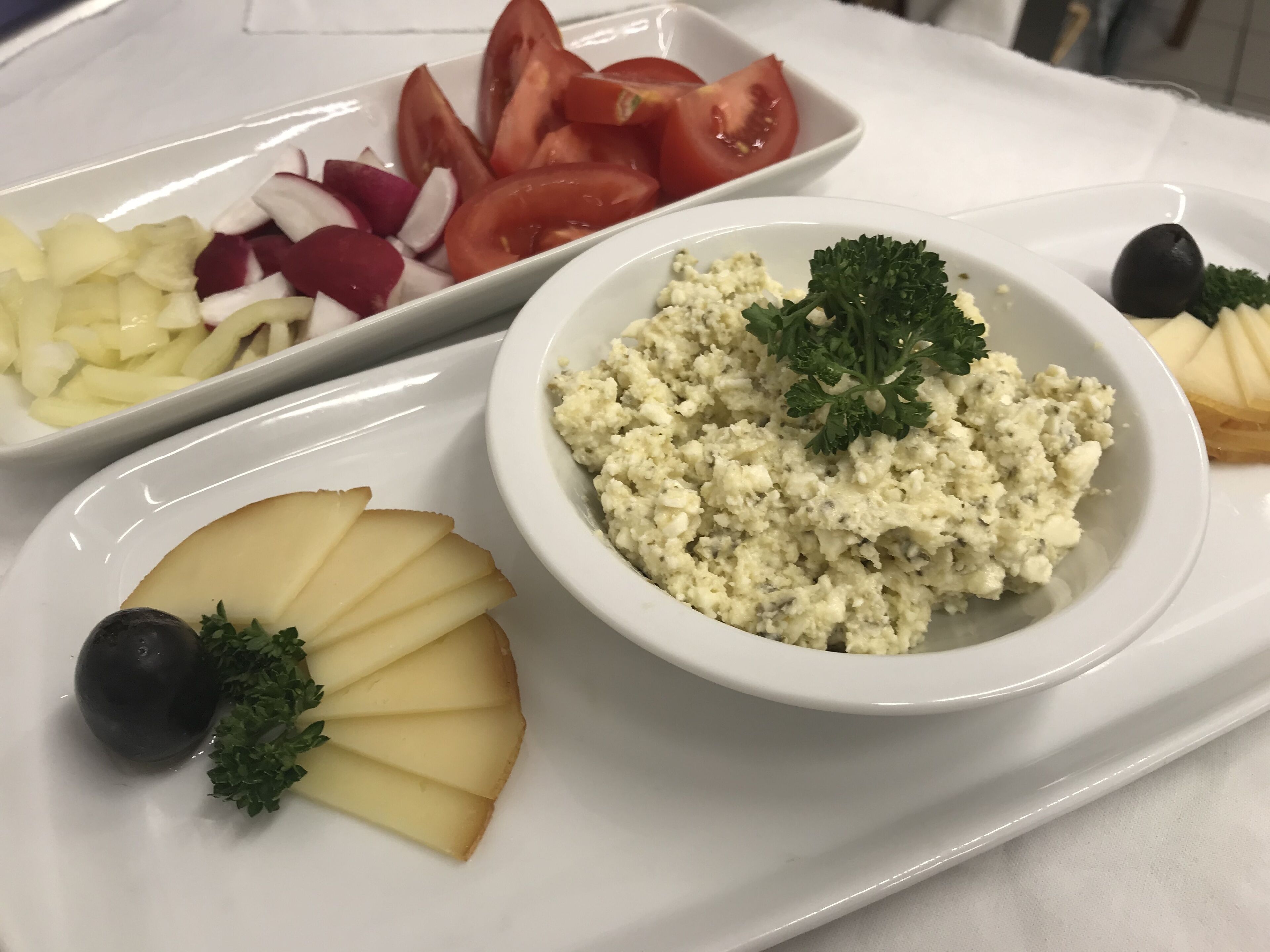 Un plateau blanc avec un saladier de salade au fromage feta, une assiette de tomates, de radis et d’oignons en tranches, et une tranche de fromage garnie d’olives et de persil.