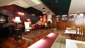 Un hall d’hôtel avec un comptoir d’accueil en bois, une moquette rouge, un fauteuil blanc et un mur orné de photographies historiques.