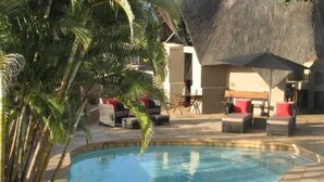 Een zwembadgedeelte met ligbedden en parasols, omgeven door palmbomen en een constructie met rieten dak.