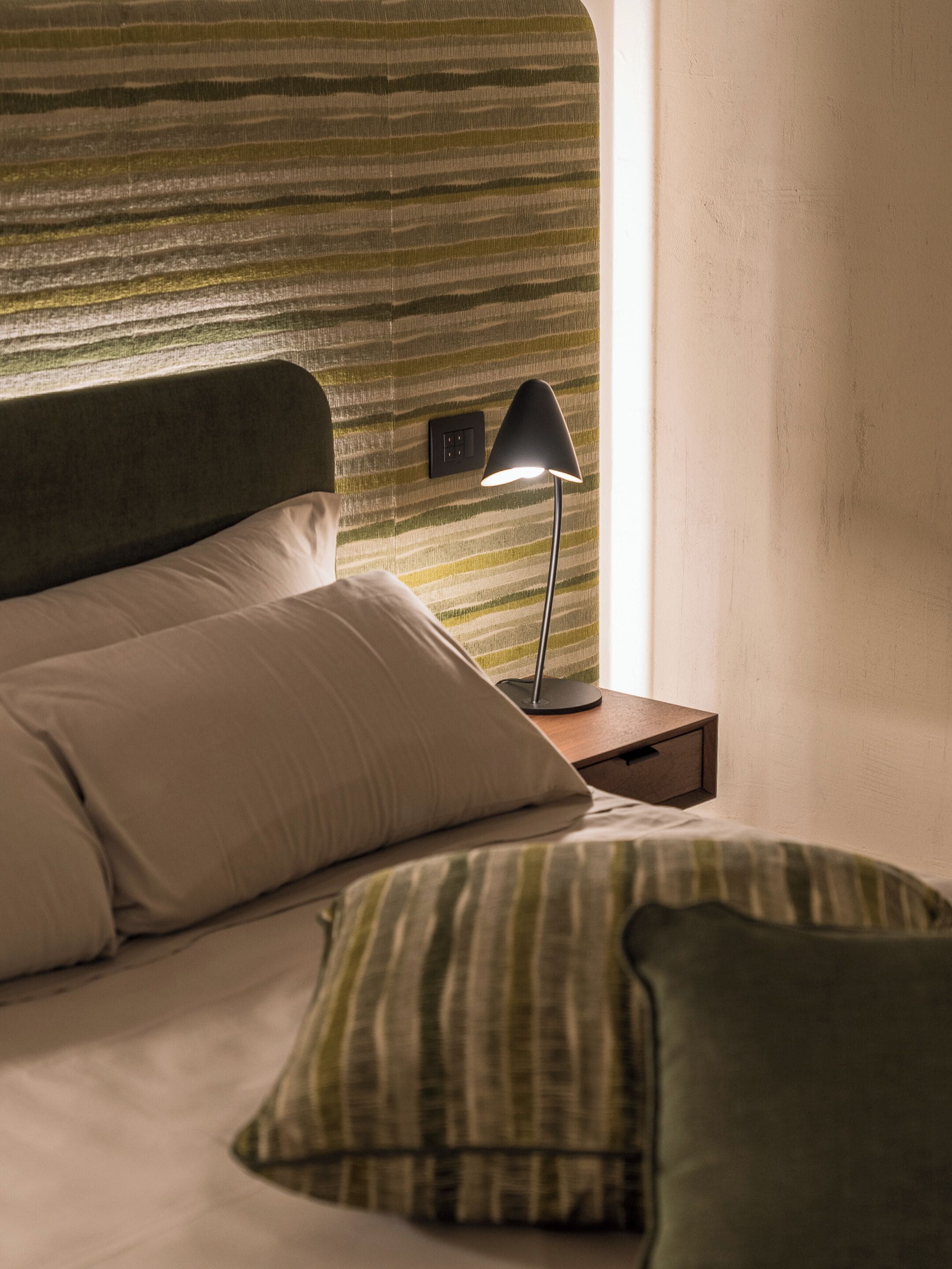 A neatly made bed with a striped headboard, a bedside lamp, and a nightstand with a power outlet.