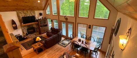 A spacious living area with a fireplace, wooden flooring, a dining table with chairs, and large windows offering a view of greenery.