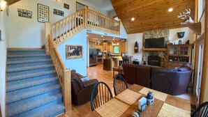 A spacious living area with a wooden staircase, a dining table with chairs, a kitchen in the background, and a stone fireplace.