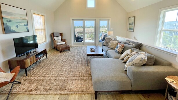 A spacious living room with a large window, a cozy sofa, a flat-screen TV, and a wooden ceiling. - Beach Nut Bungalow:  Two bedroom home in Pamlico Point (Ocracoke)
