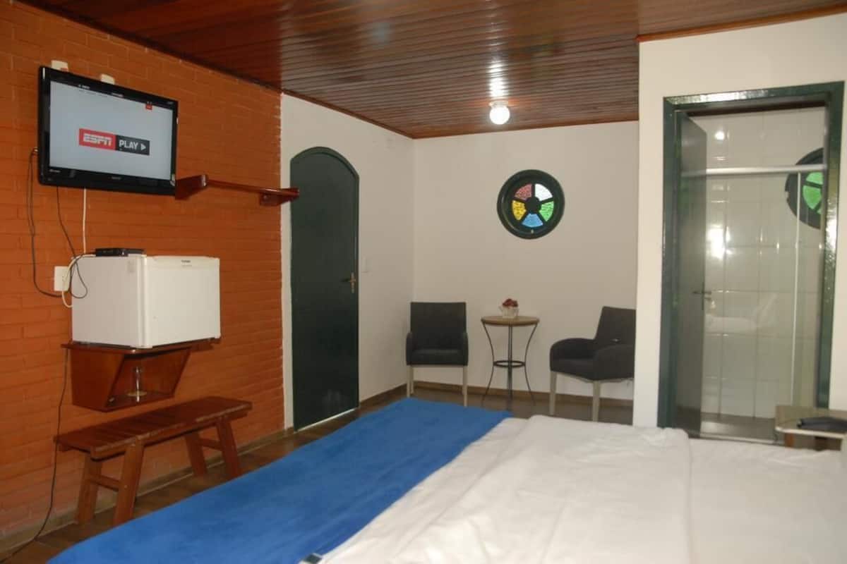 Habitación de hotel con cama, televisor montado en la pared, una pequeña mesa con dos sillas y una puerta que da a un área de ducha.