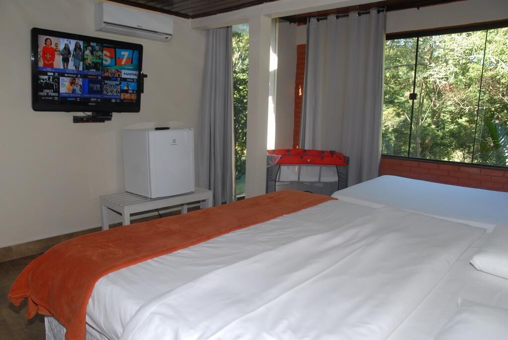 A hotel room with a bed, a TV mounted on the wall, a small refrigerator, and a window with curtains.