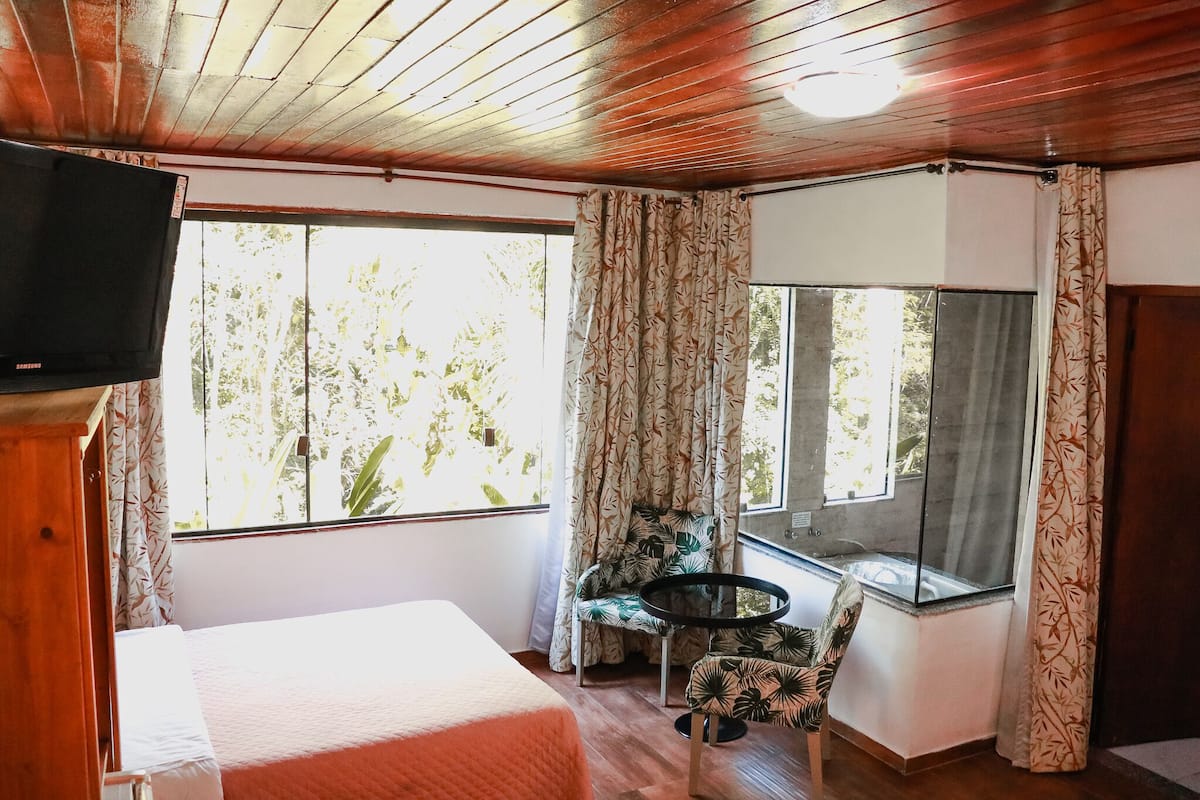 Una habitación con cama, televisión, ventana con cortinas, una silla y una mesita.
