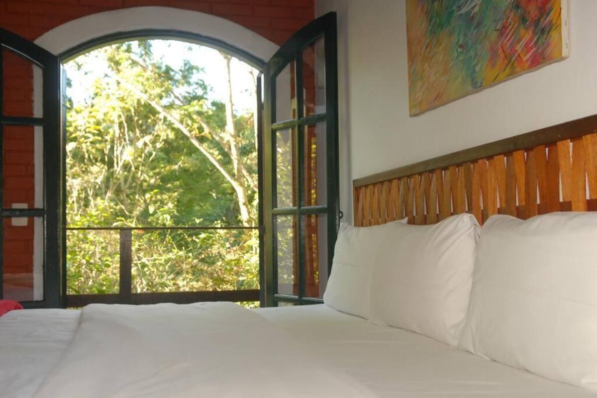 Una cama con sábanas blancas, cabecera de madera y un gran ventanal con la puerta abierta que muestra vistas de árboles.