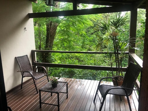 A balcony with two wicker chairs and a small table, overlooking a lush green forest. -  Pousada Mirante do Penedo (Itatiaia)