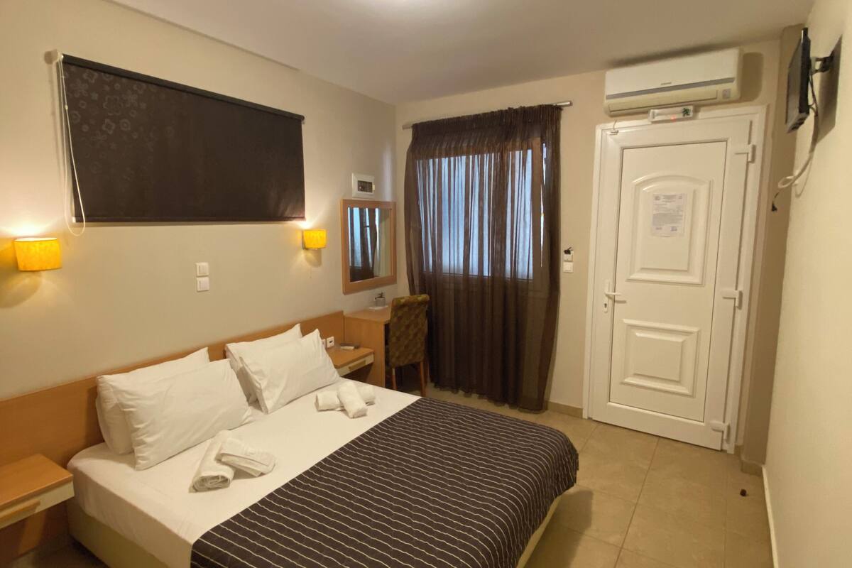 Una habitación de hotel con cama, lámparas de noche, escritorio, silla, armario y aire acondicionado.