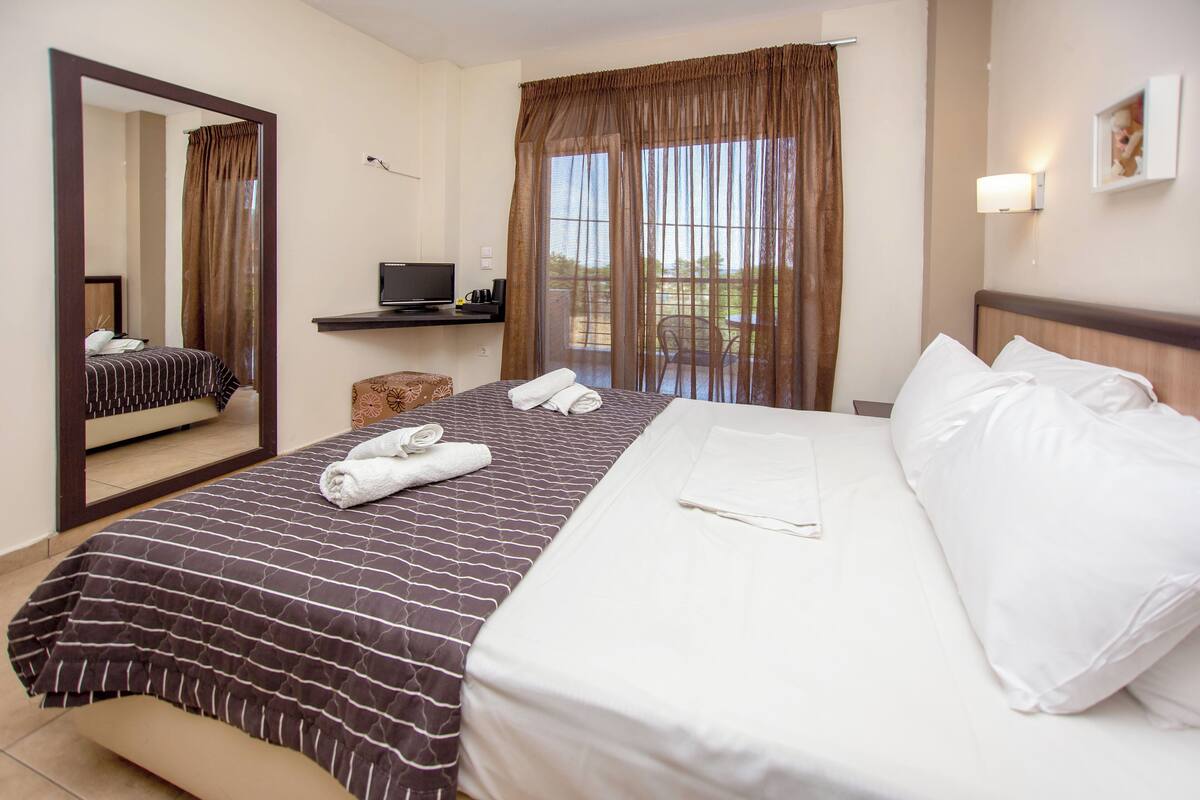 Habitación de hotel con una cama grande, televisión, una mesita y un balcón con cortinas.