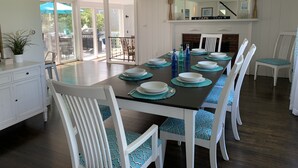 A dining room with a long table set for six, white chairs with blue patterned cushions, and a fireplace with a 'CAPE COD' sign above it.