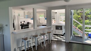 A modern kitchen with white cabinetry, a stainless steel refrigerator, a microwave, and a stove. There are three white bar stools and a large window providing a view of the outdoors.