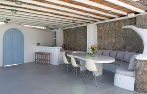 A covered outdoor dining area with a stone wall, a blue door, and a white table with chairs. - Villa Isabella (Mykonos)