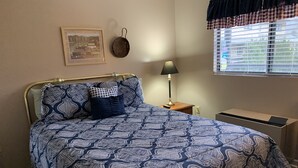 Una camera da letto con una coperta a motivi, un quadro appeso al muro, una lampada e una finestra con persiane.