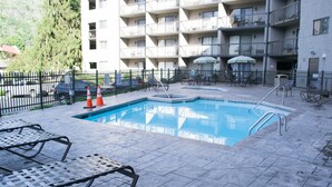 Una piscina con lettini e ombrelloni, circondata da un'area recintata con un edificio a più piani sullo sfondo.