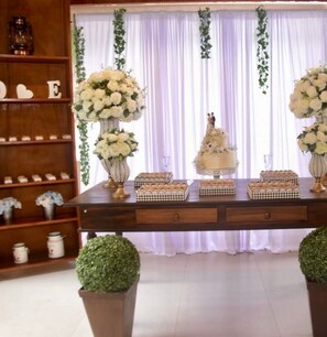 A decorated table with floral arrangements, a cake, and checkered boxes, set against a backdrop with white curtains and greenery.