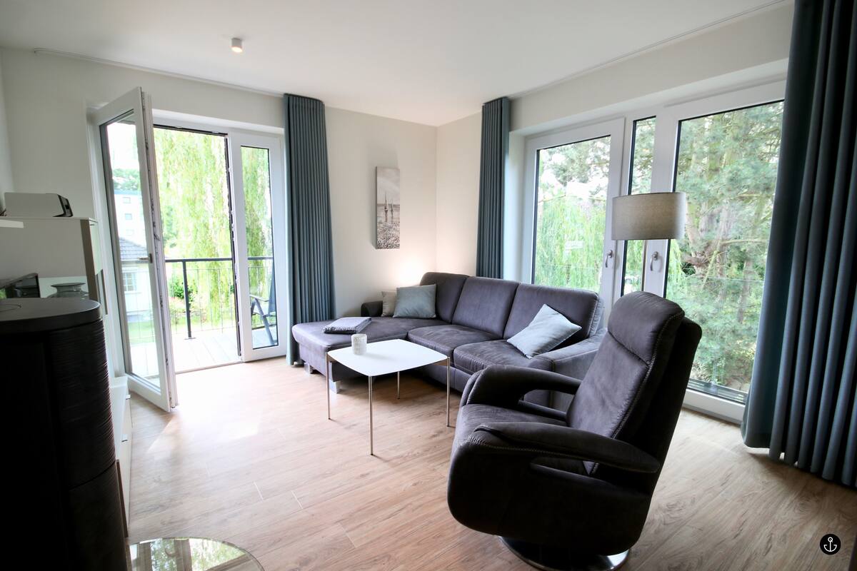 Un salon moderne avec un canapé d’angle, une table basse et une lampe. Une grande fenêtre, ornée de rideaux, et une porte donnant sur un espace extérieur complètent la pièce.