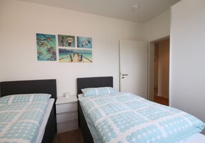 A bedroom with two beds, a nightstand, a dresser, and wall art depicting a beach scene. - Kapitänshaus 4 (Kellenhusen)