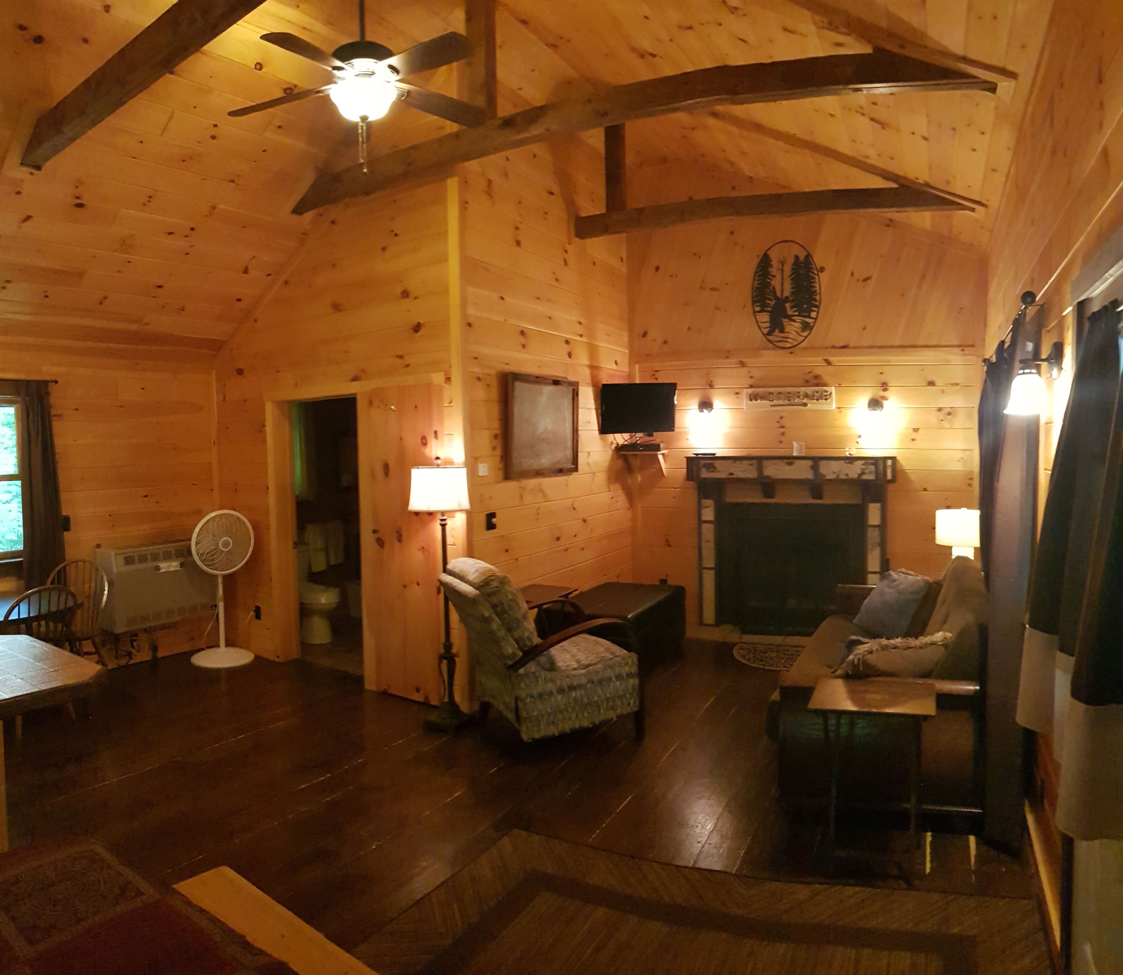 A cozy cabin interior with a wooden ceiling, a fireplace, a television, and a dining area.