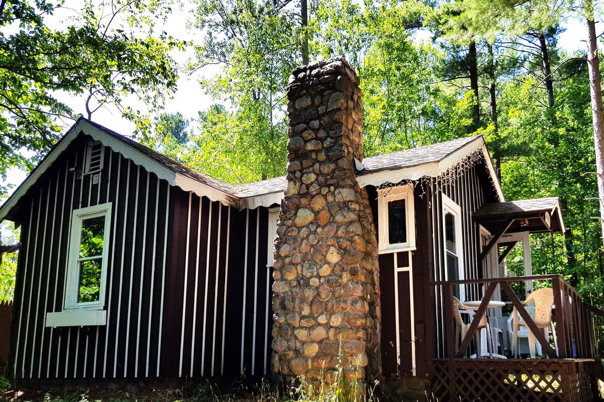 A rustic cabin with a stone chimney, wooden siding, and a porch with chairs.
