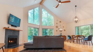 A spacious living room with a fireplace, a large sofa, a dining area, and large windows offering a view of the outdoors.