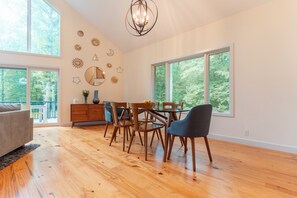 A spacious dining area with wooden flooring, a dining table with chairs, and large windows offering a view of trees.