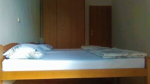 Ein Einzelbett mit Holzrahmen, einer weißen Matratze und zwei Kissen. Dazu steht eine Holzgarderobe mit einer Tür rechts neben dem Bett.