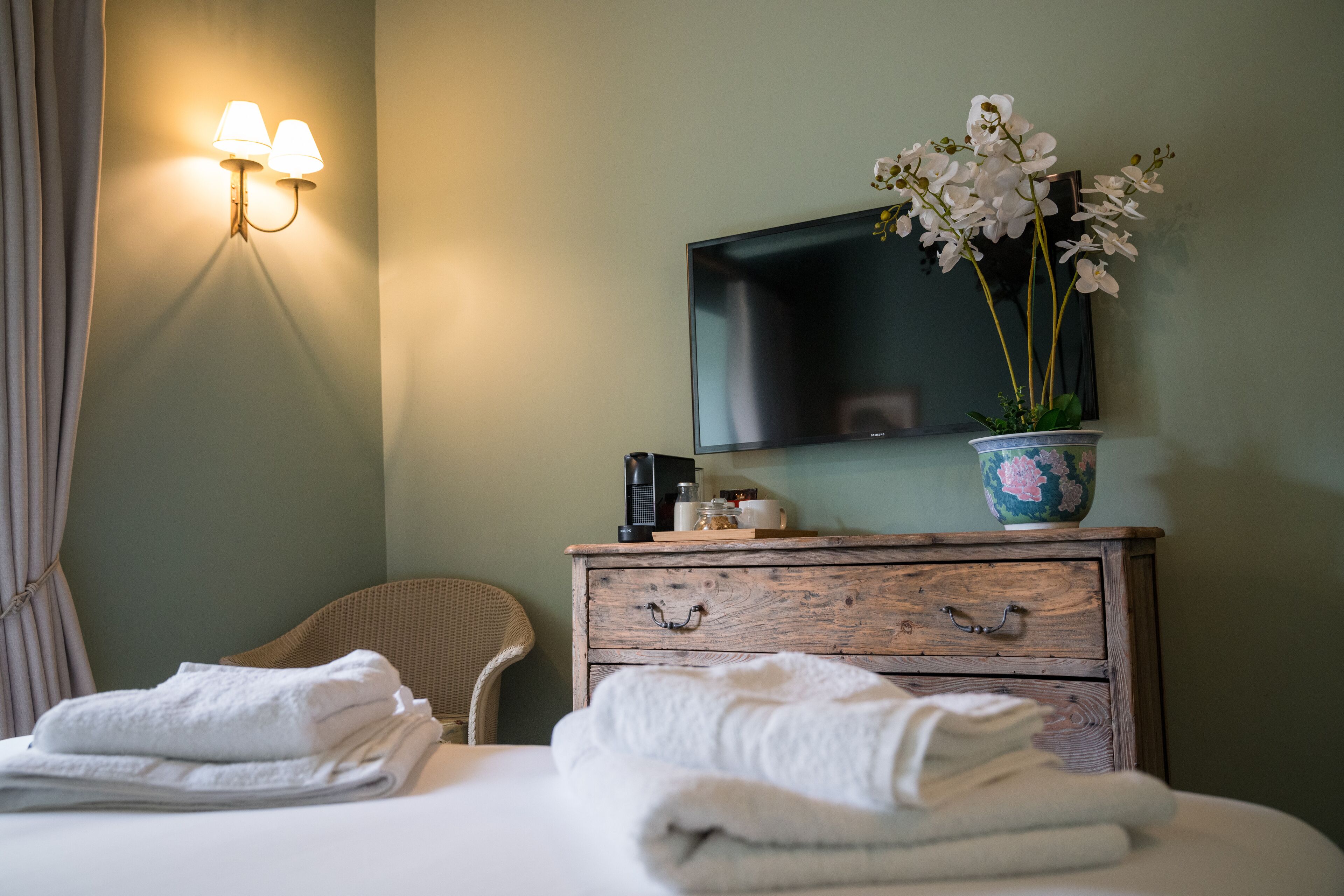 A hotel room with a bed, a wooden dresser, a television, a vase with flowers, and a wall-mounted light fixture.