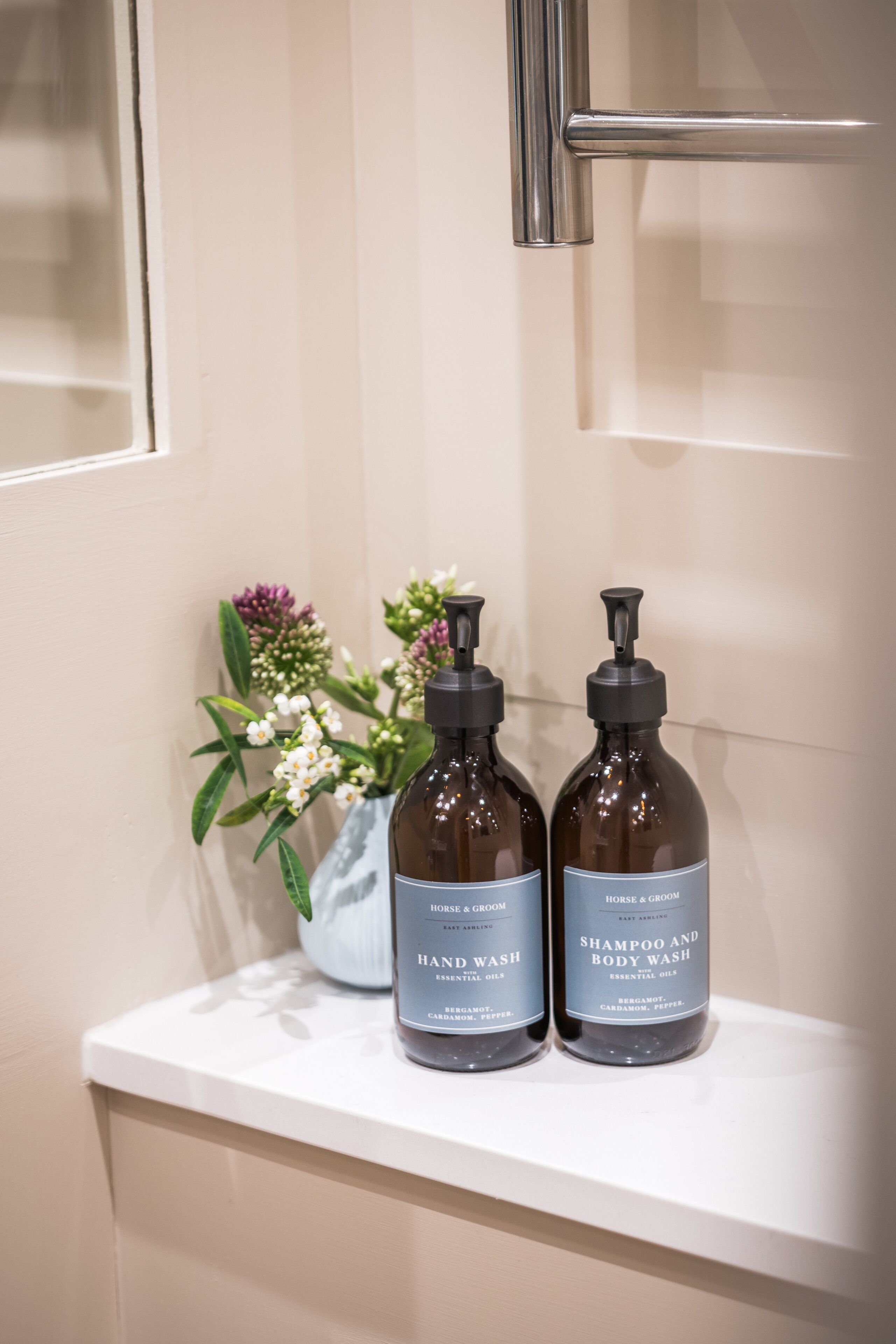 A bathroom with a white countertop, two brown bottles labeled "Hand Wash" and "Shampoo and Body Wash," and a vase with flowers.