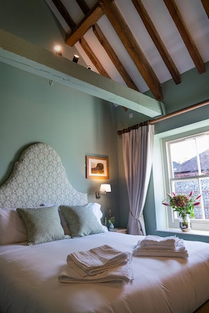 Un lit bien fait, avec des serviettes pliées, une tête de lit ornée d’un motif floral, une lampe de chevet et une fenêtre avec des rideaux.