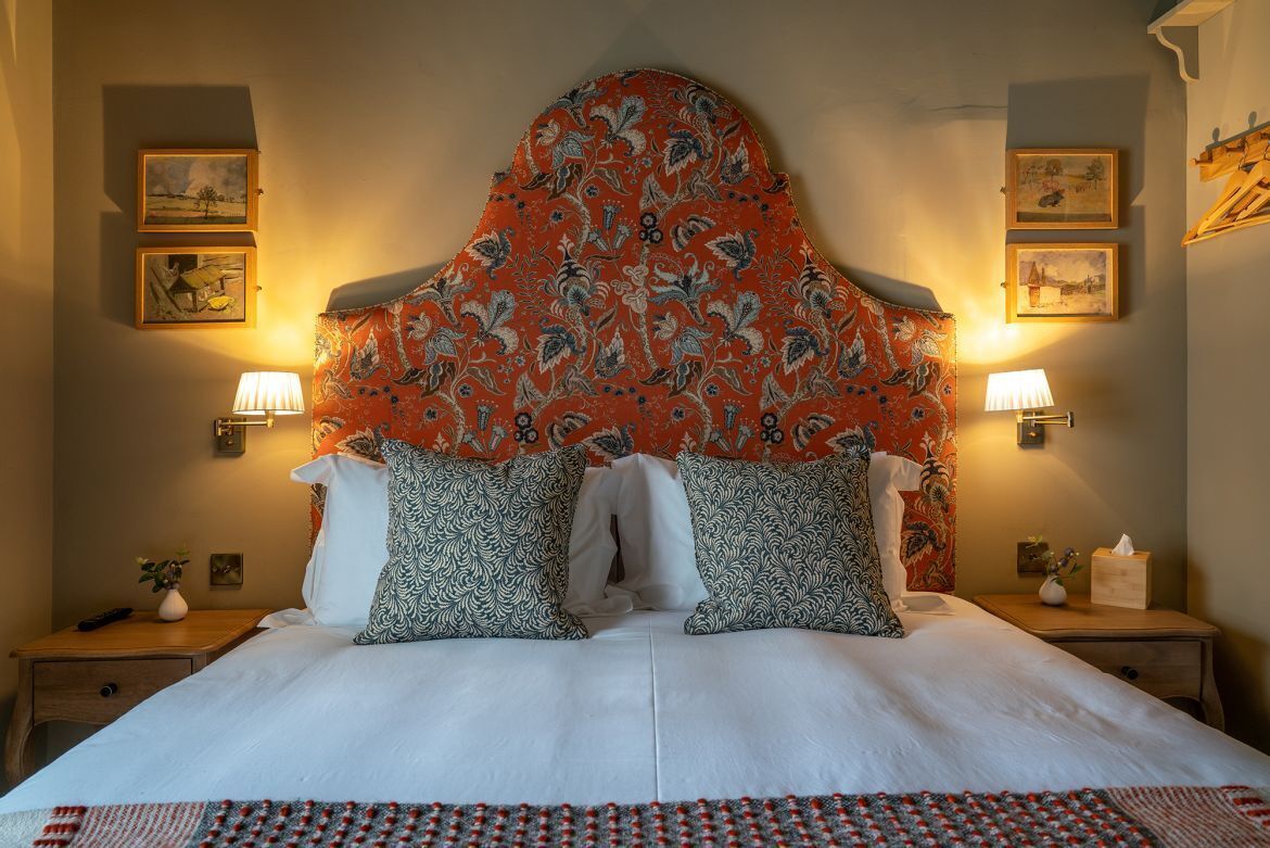 Uma cama de casal com cabeceira estampada, duas luminárias de mesa e quadros nas paredes.