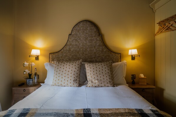 A neatly made bed with a patterned headboard, two bedside lamps, and a nightstand with a vase of flowers and a bottle. - The Horse and Groom (Chichester)