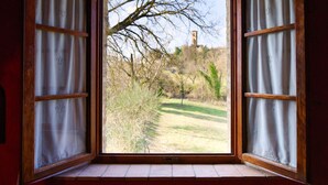 Una ventana abierta con marcos de madera y cortinas blancas, que ofrece vistas a un paisaje cubierto de césped con árboles y un edificio lejano.