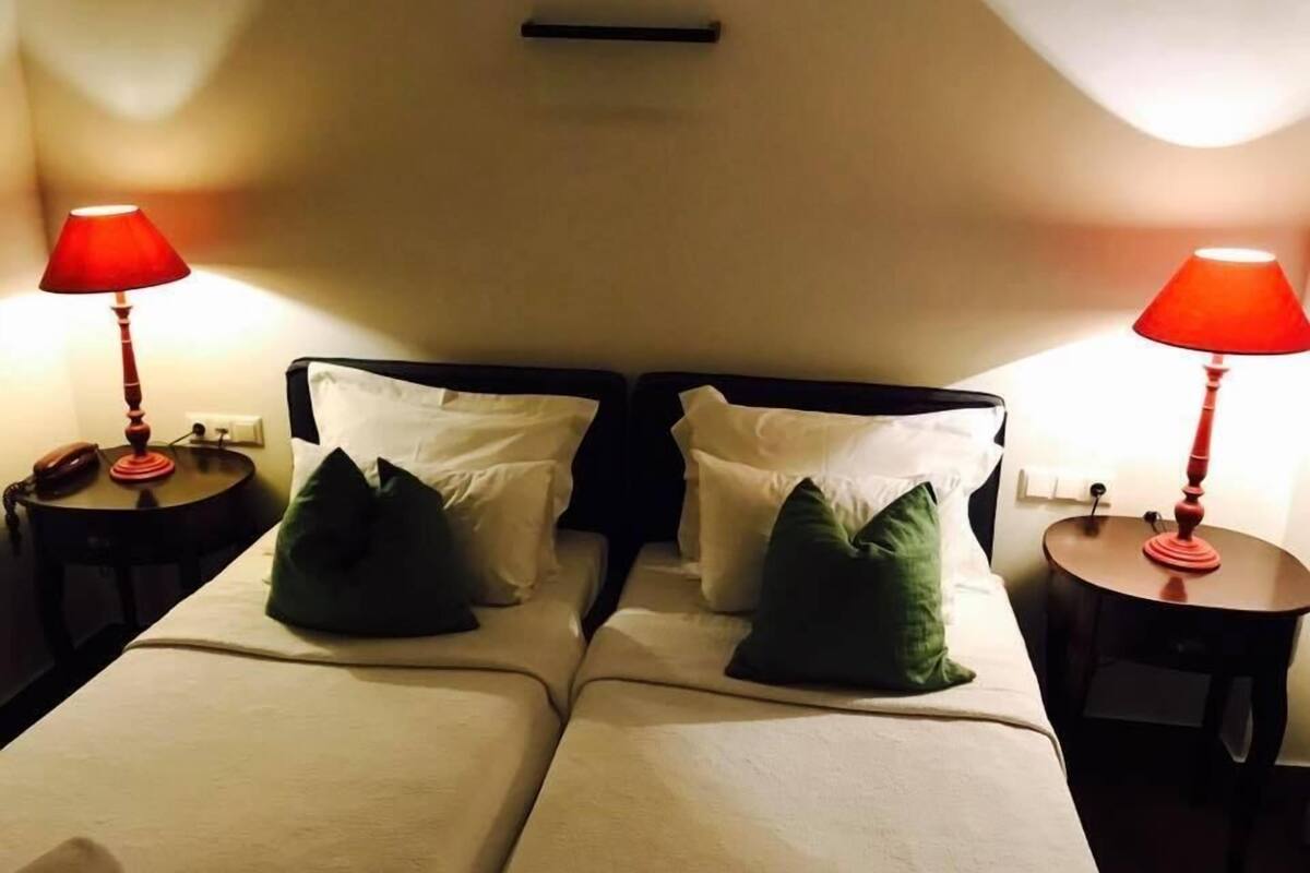 Habitación de hotel con dos camas, cada una con ropa blanca y almohadas verdes, flanqueadas por mesitas de noche con lámparas rojas.