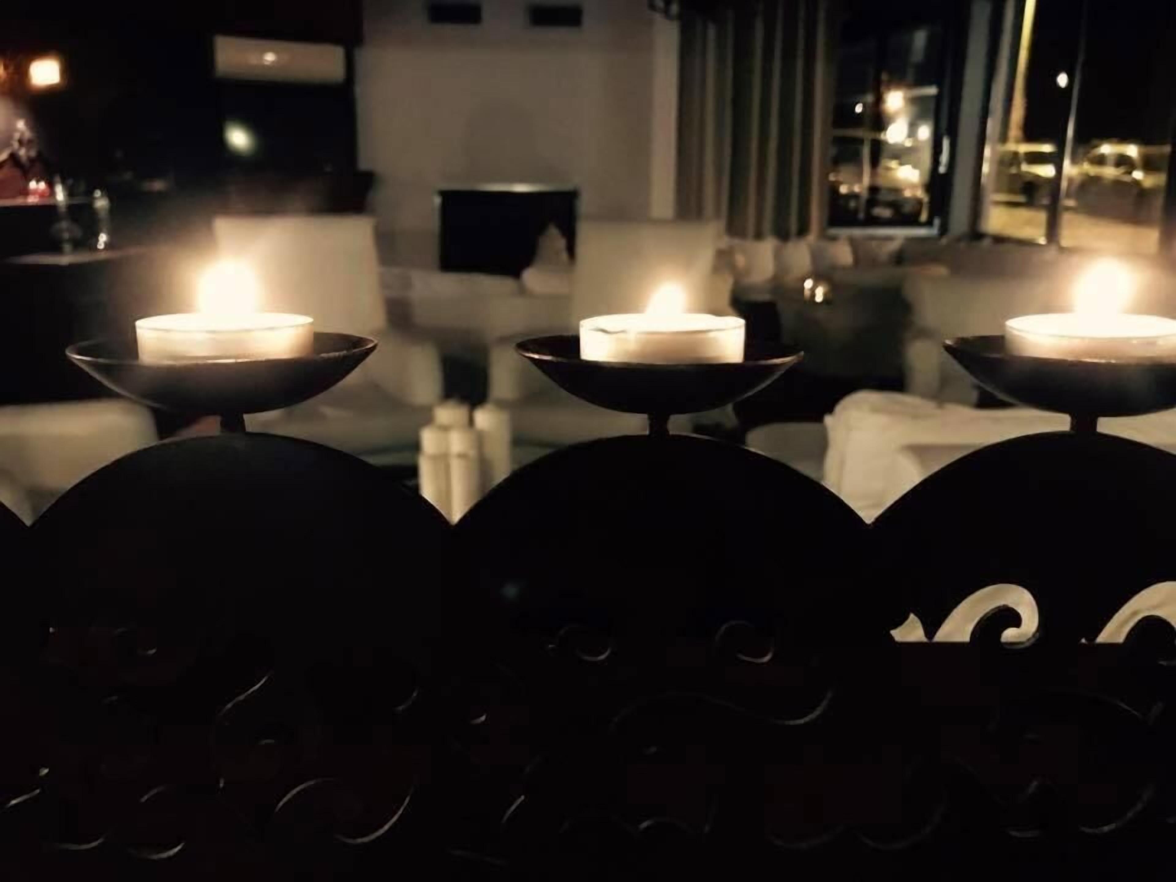 A dimly lit outdoor area with three lit candles on stands, a white wall, and a window in the background.