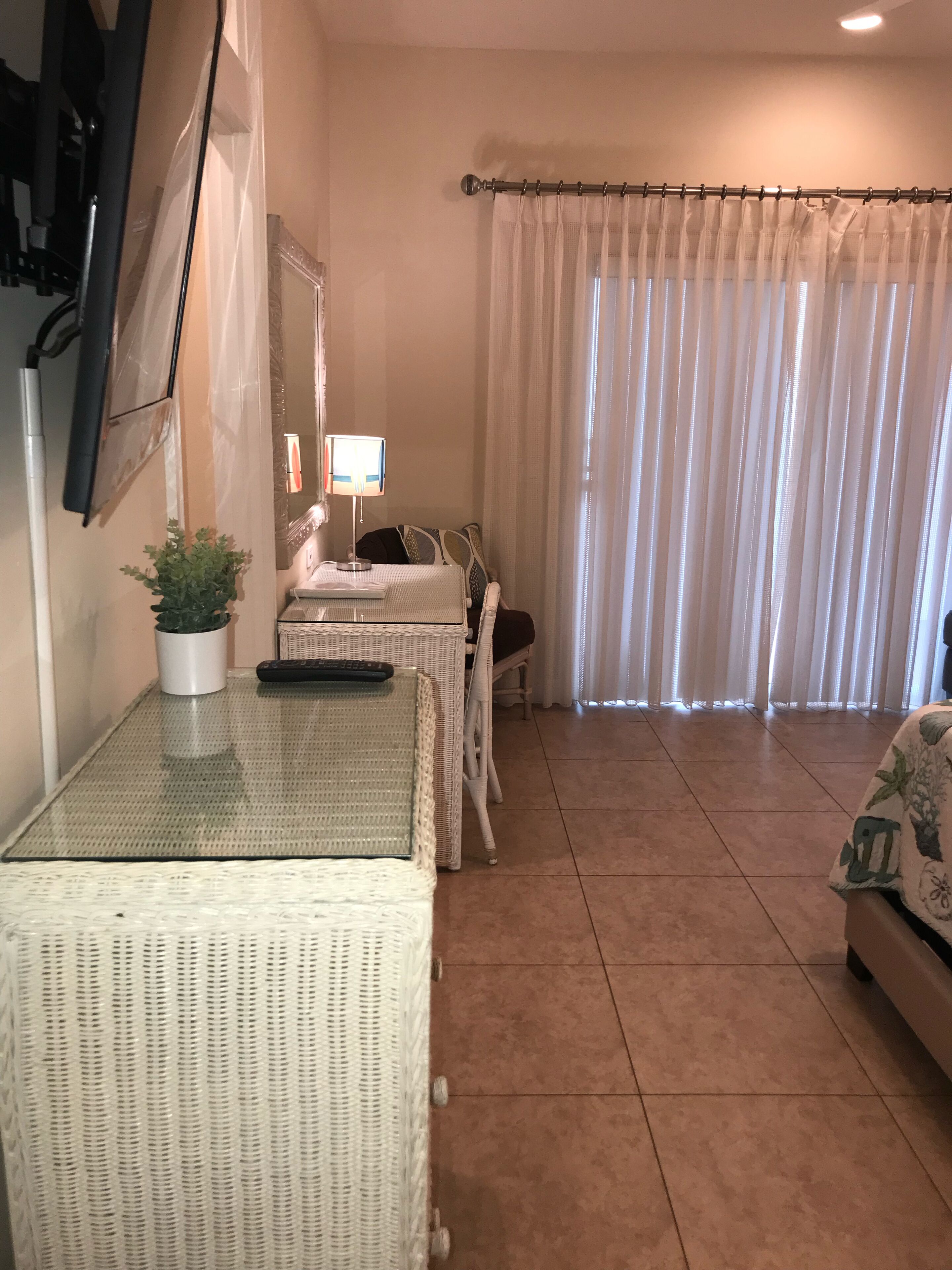 A room with a glass-top table, a potted plant, a lamp, a bed with a white headboard, and a curtain.